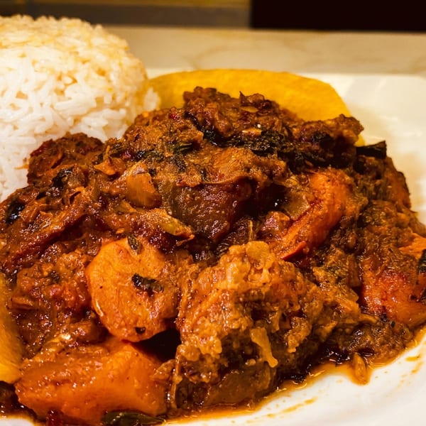 A plate of stewed meat and vegetables with white rice, fried plantain, and a blue bowl of soup.